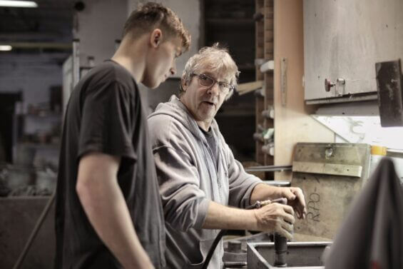 Older teacher explains to student how to operate workshop tool.