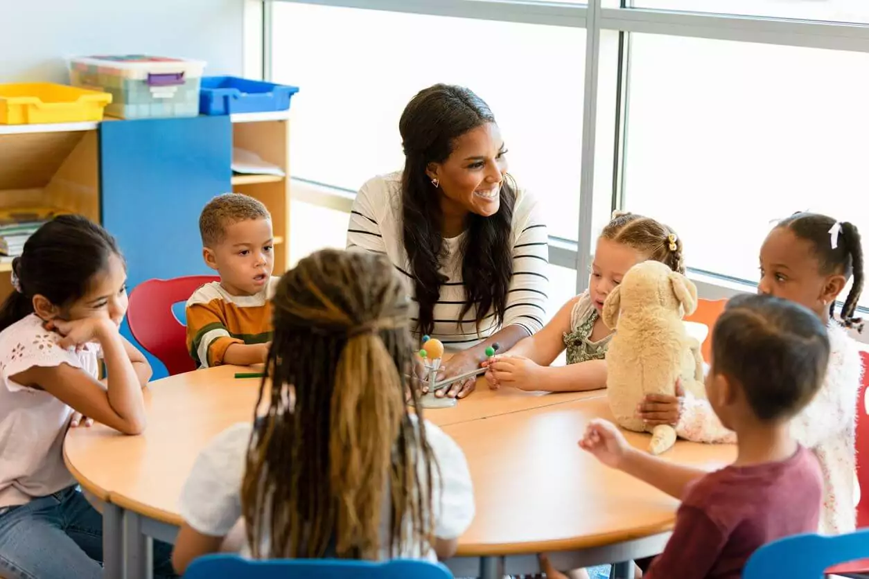 Smiling teacher sits at a small round table with students discussing the solar system.