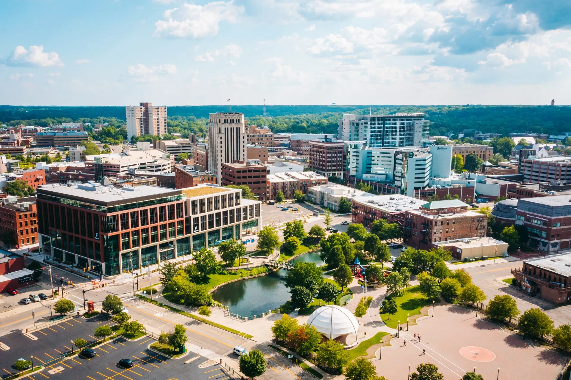 An aerial view of Kalamazoo shows a park with mature trees and a pond set amongst the buildings.