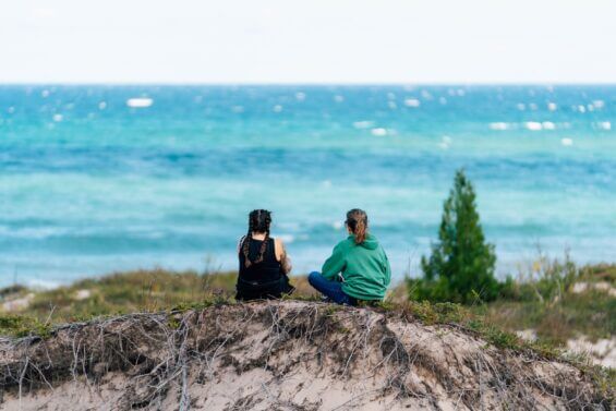 Two women sit on a sandy hill at Thompson's Harbor State Park.