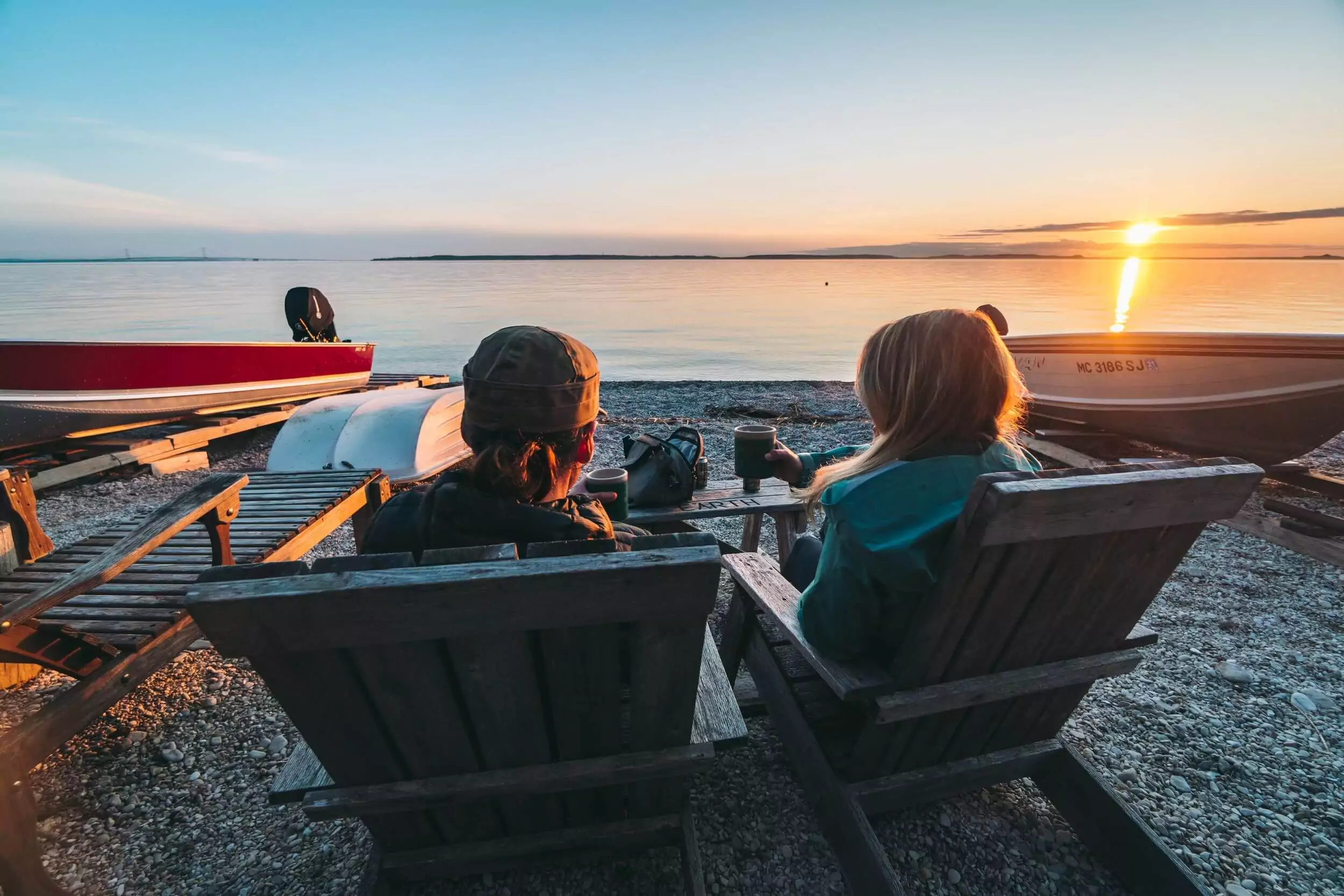Two people sit in wooden beach chairs at sunset.